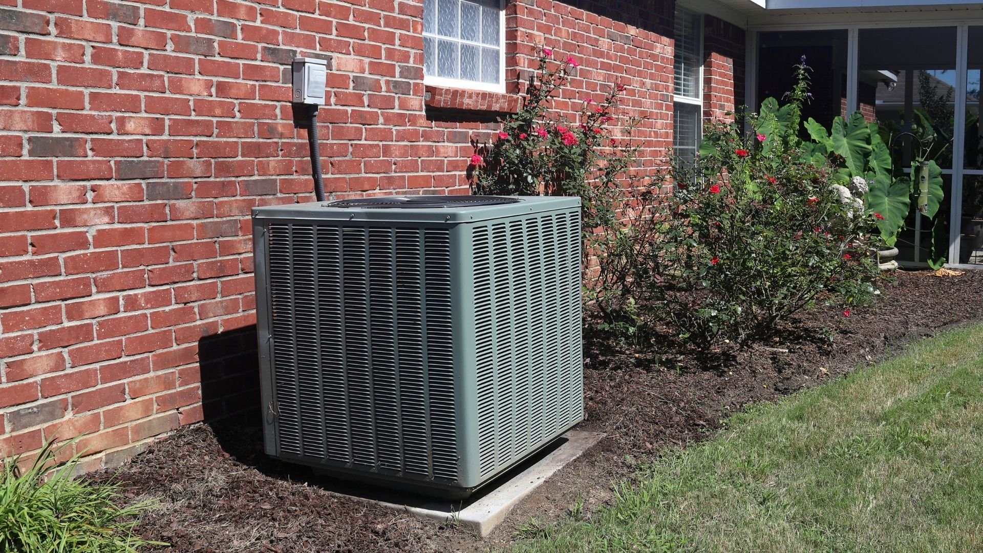 Outdoor AC unit against red brick house with flowering shrubs nearby